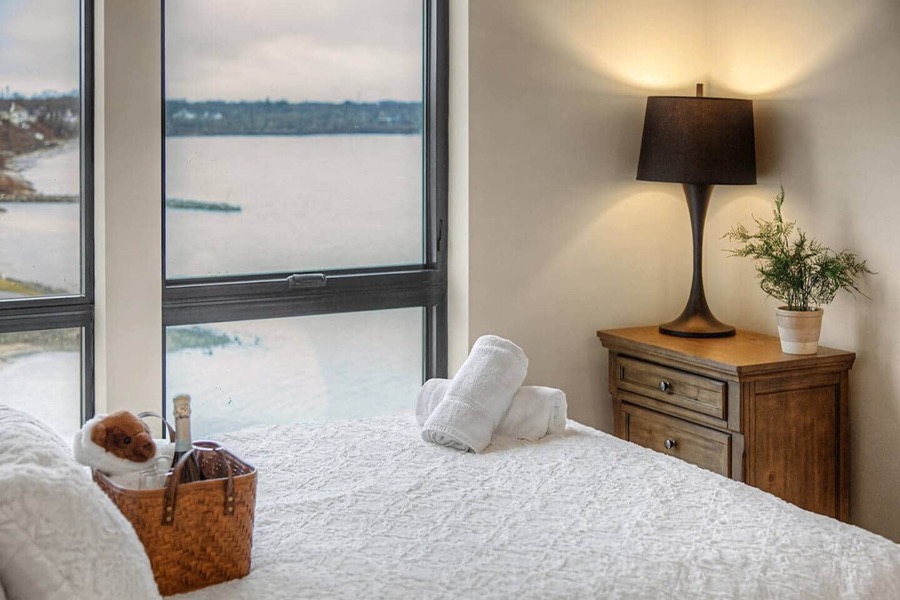 A lake-view bedroom — white bedding, a wooden dresser with lamp, and Muskegon Lake through the corner window