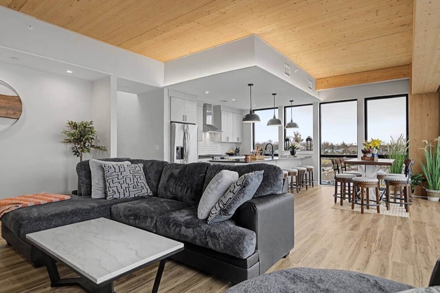 Open-plan living room of Residence 601 with a plush charcoal sectional, wood-plank ceiling, and floor-to-ceiling windows framing Muskegon Lake