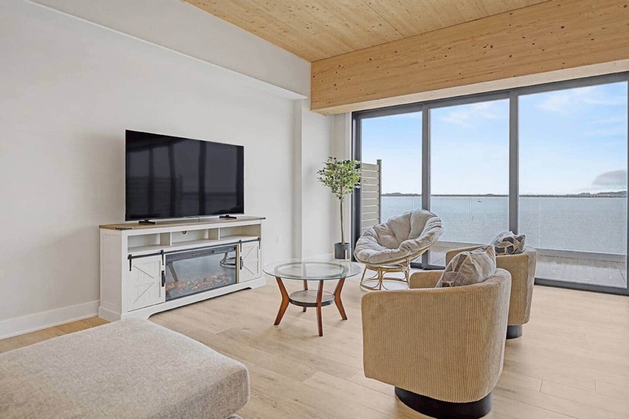 Living area with the TV atop a white barn-door console with electric fireplace, cream swivel chairs, and Muskegon Lake through the glass