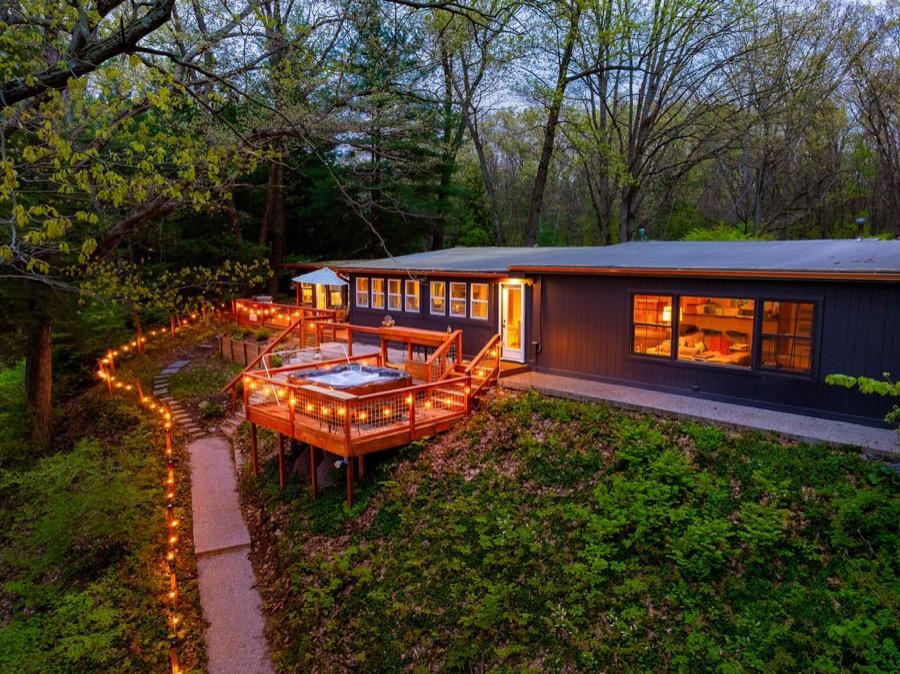 Fire & Water's dark-sided riverside cabin at dusk, string lights tracing the path down to a deck with hot tub above the Kalamazoo River