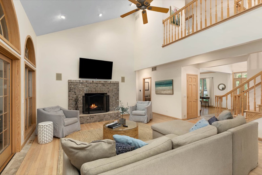 The two-story great room: brick fireplace with mounted TV, plush seating, and the balcony landing above