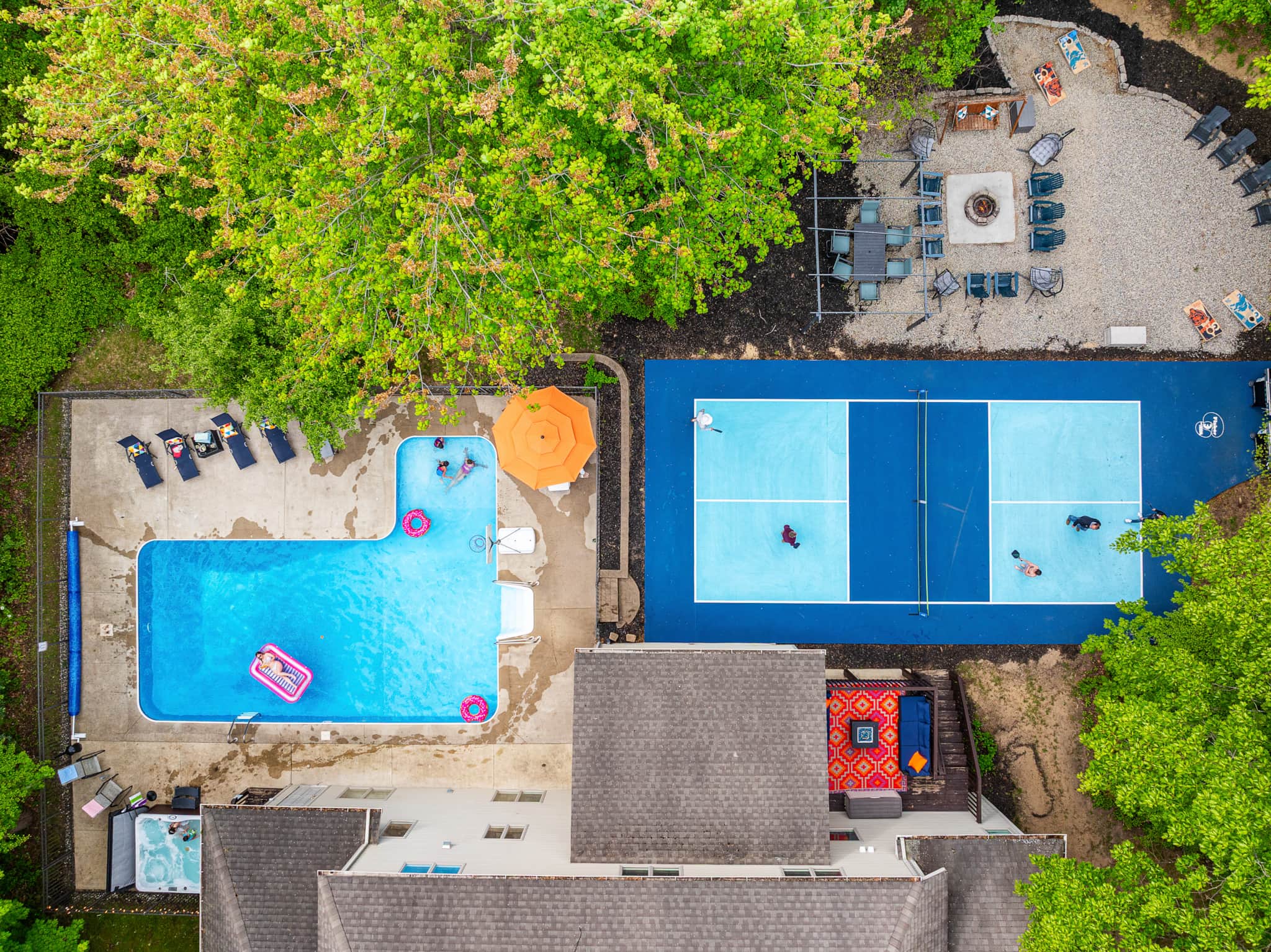 Aerial view over Swimming in Amenities in Holland — an L-shaped inground pool with floats, a blue pickleball court mid-game, and a gravel fire-pit lounge tucked into the trees