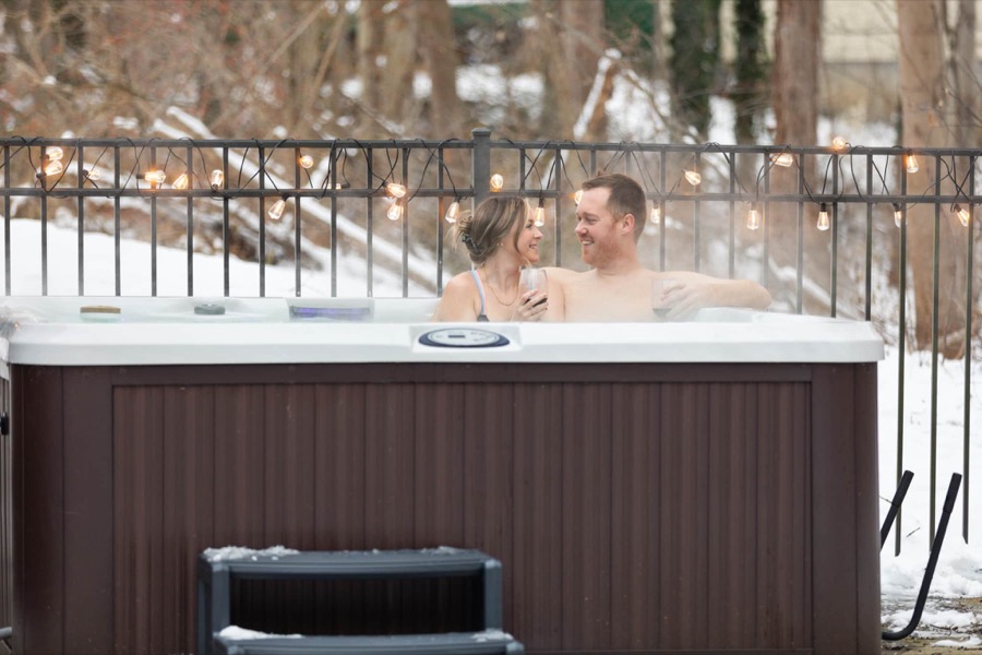 A couple enjoying the steaming hot tub on a snowy winter day, string lights glowing along the fence — the spa runs year-round