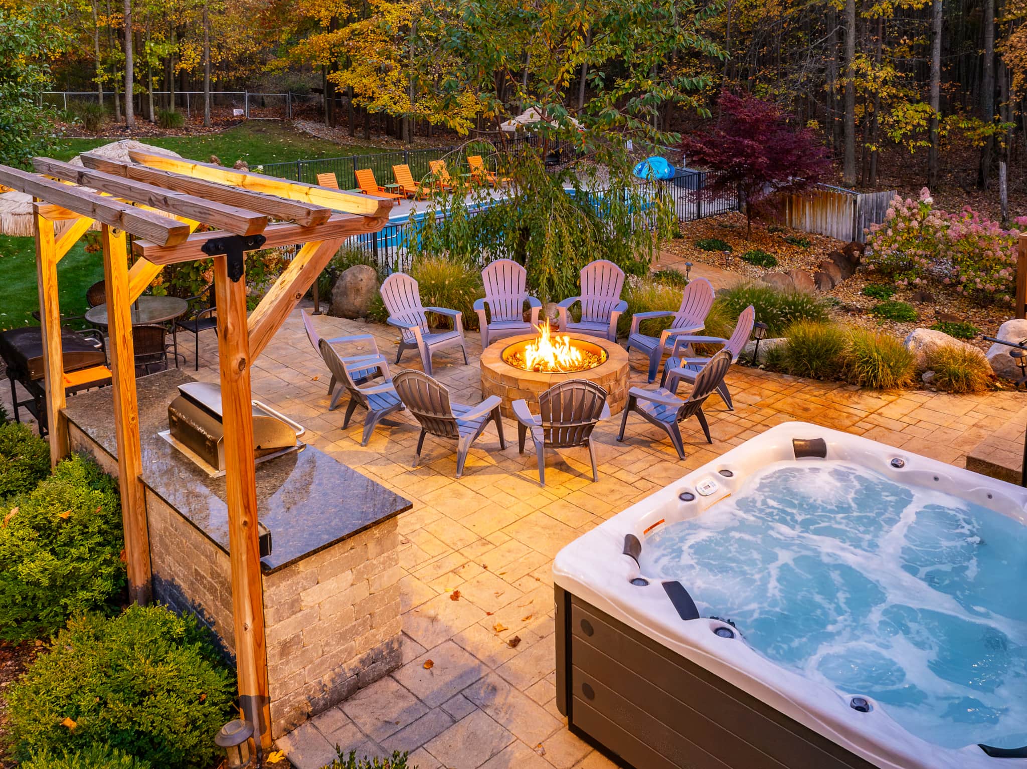 Backyard of Swimming in the Sunset at dusk — a bubbling hot tub beside a stone fire pit ringed with Adirondack chairs, a pergola-covered outdoor kitchen, and the heated pool glowing beyond