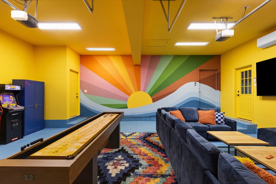 Game room at Swimming in the Sunset with a sunset mural covering the wall, shuffleboard table, arcade cabinet, and a navy sectional sofa