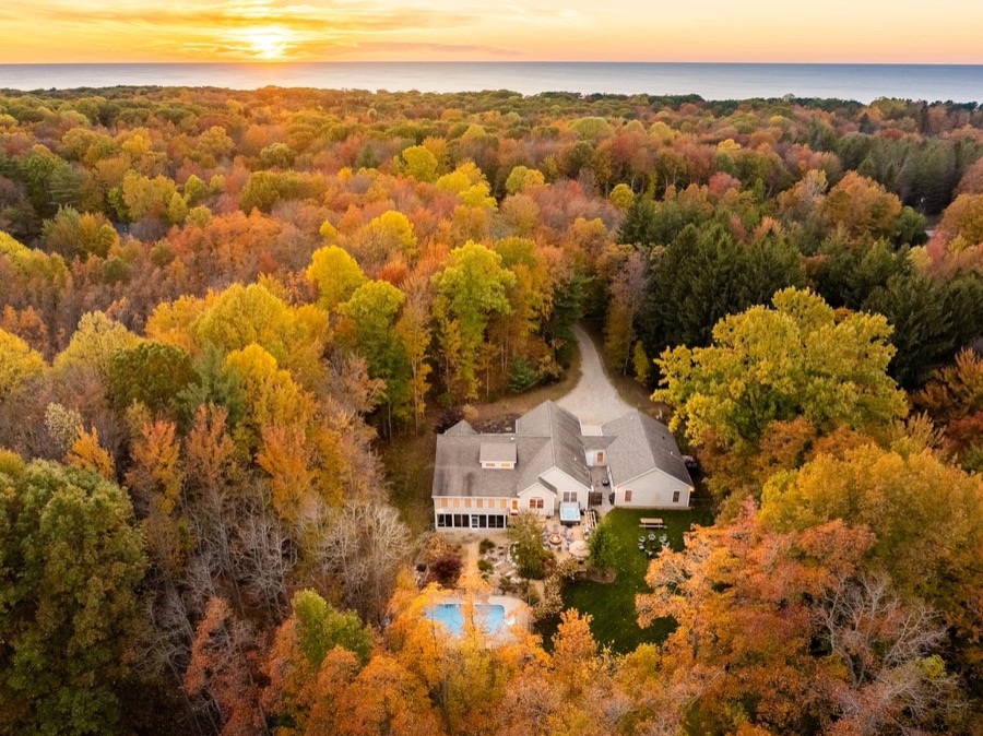 Aerial autumn view of the home tucked into private wooded acreage, pool below and Lake Michigan glowing on the sunset horizon
