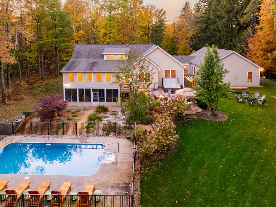 Aerial view of the estate at golden hour: the main house and apartment wing, hot tub deck, fire-pit patio, Adirondack circle on the lawn, and fenced pool with orange loungers