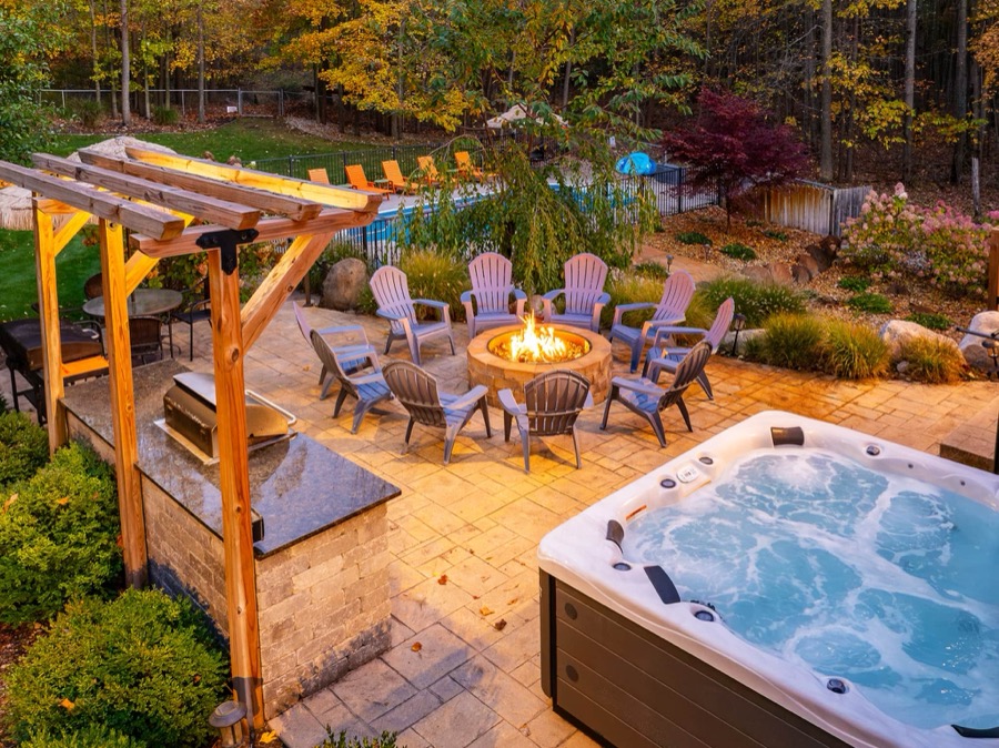 Backyard of Swimming in the Sunset at dusk — a bubbling hot tub beside a stone fire pit ringed with Adirondack chairs, with a pergola-covered outdoor kitchen and the pool beyond